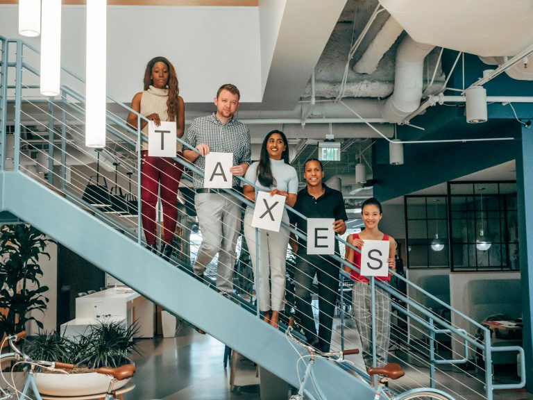 Multicultural business team holding 'Taxes' signs on a modern office staircase, symbolizing collaboration.