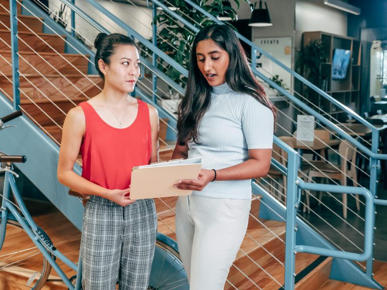 Two young women engage in a discussion in a modern office setting, promoting teamwork and diversity.